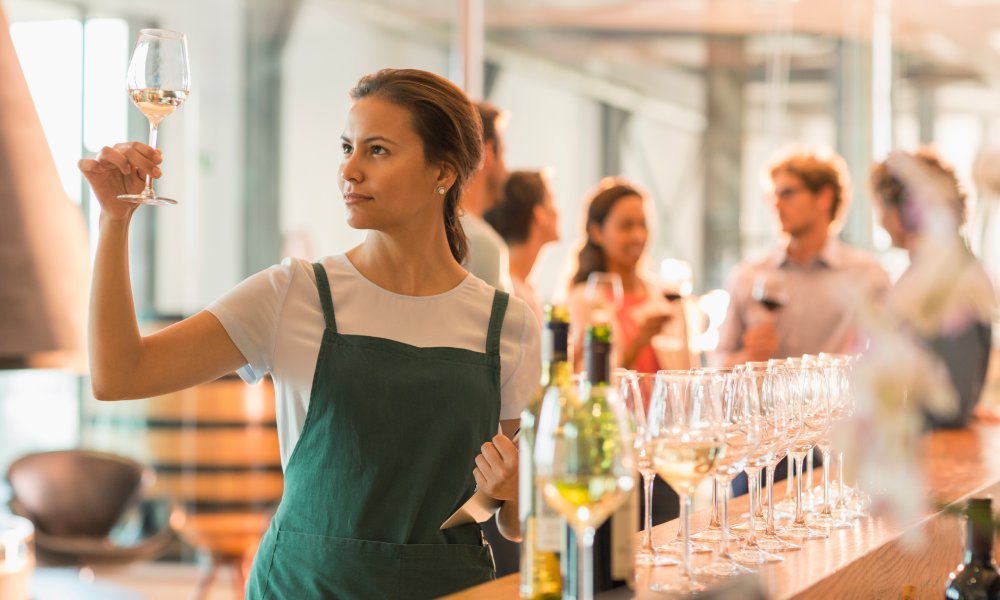 Female wine tasting room worker behind a bar and holding a glass of white wine up to examine it. Guests are laughing in the background.
