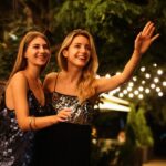 Two young blonde women in sparkly cocktail dresses standing outside on a city street laughing and hailing a cab.