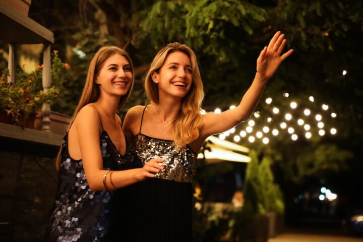 Two young blonde women in sparkly cocktail dresses standing outside on a city street laughing and hailing a cab.