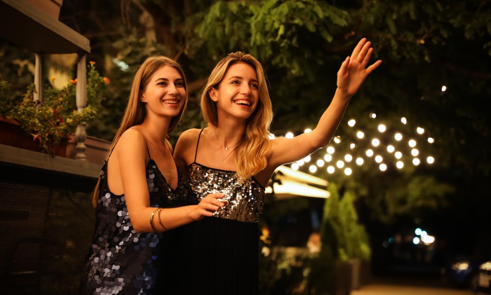 Two young blonde women in sparkly cocktail dresses standing outside on a city street laughing and hailing a cab.